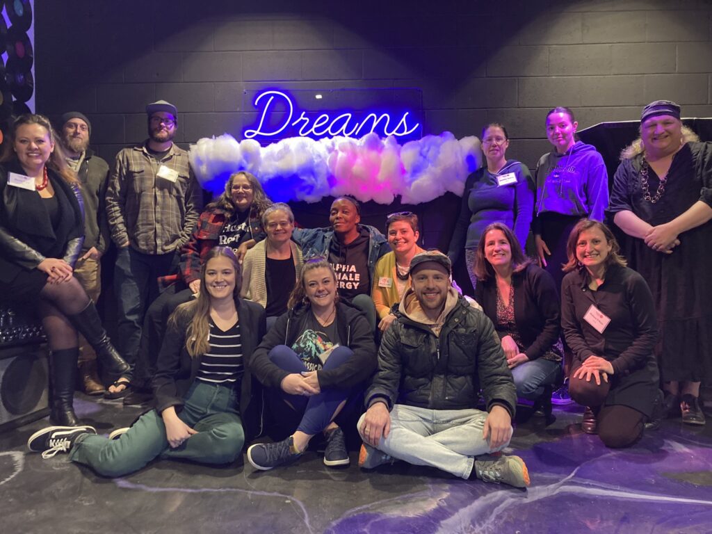 A group of people pose indoors in front of a blue neon sign that reads "Dreams," surrounded by faux clouds. They are casually dressed, smiling, and seated or standing in a relaxed manner, suggesting a friendly gathering or event.
