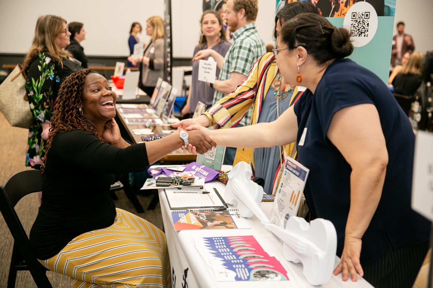 A woman sitting at a table at a job fair enthusiastically shakes hands with a visitor. The table is filled with pamphlets and flyers. In the background, other people are engaged in conversations. The atmosphere is lively and friendly.