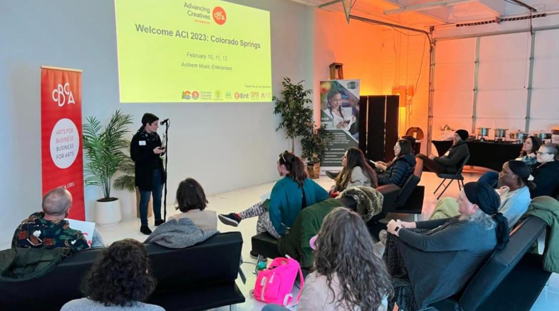 A group of people seated in a room listens to a speaker at the front. A screen displays "Welcome ACI 2023: Colorado Springs" and details about the event. Plants and banners are placed around the room.