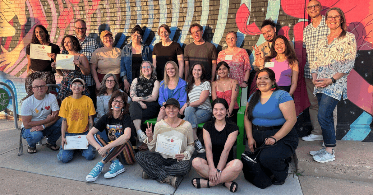 A diverse group of people poses together outside in front of a colorful mural, celebrating the Colorado Arts nonprofit's success. Some hold certificates, and everyone is smiling. Casually dressed, a few make peace signs. The atmosphere is joyful and focused on future priorities for 2025.