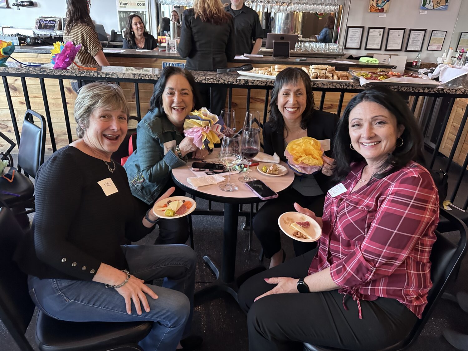 Four women sit around a small table at a restaurant, smiling at the camera. They have plates of food and glasses of wine, with two women holding flower bouquets. Name tags are visible on their shirts.