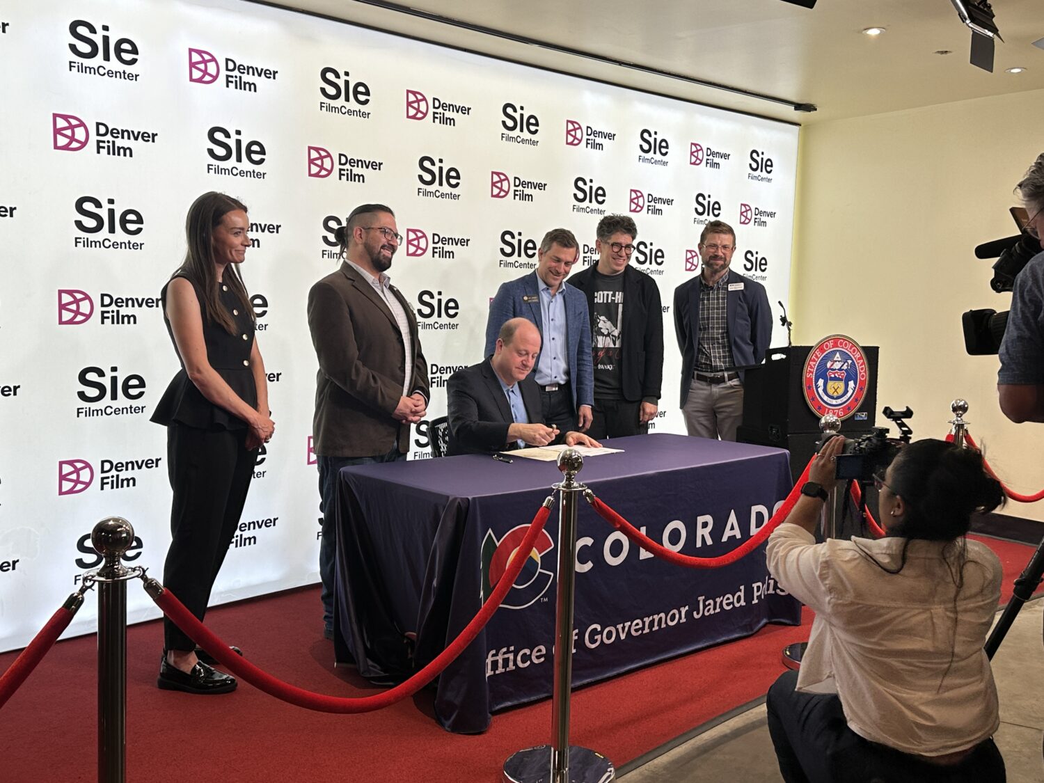 A group of people stands behind a seated man signing a document at a table with “Colorado Office of Governor Jared Polis” written on the front, with photographers capturing the moment at an event with Denver Film Center banners.