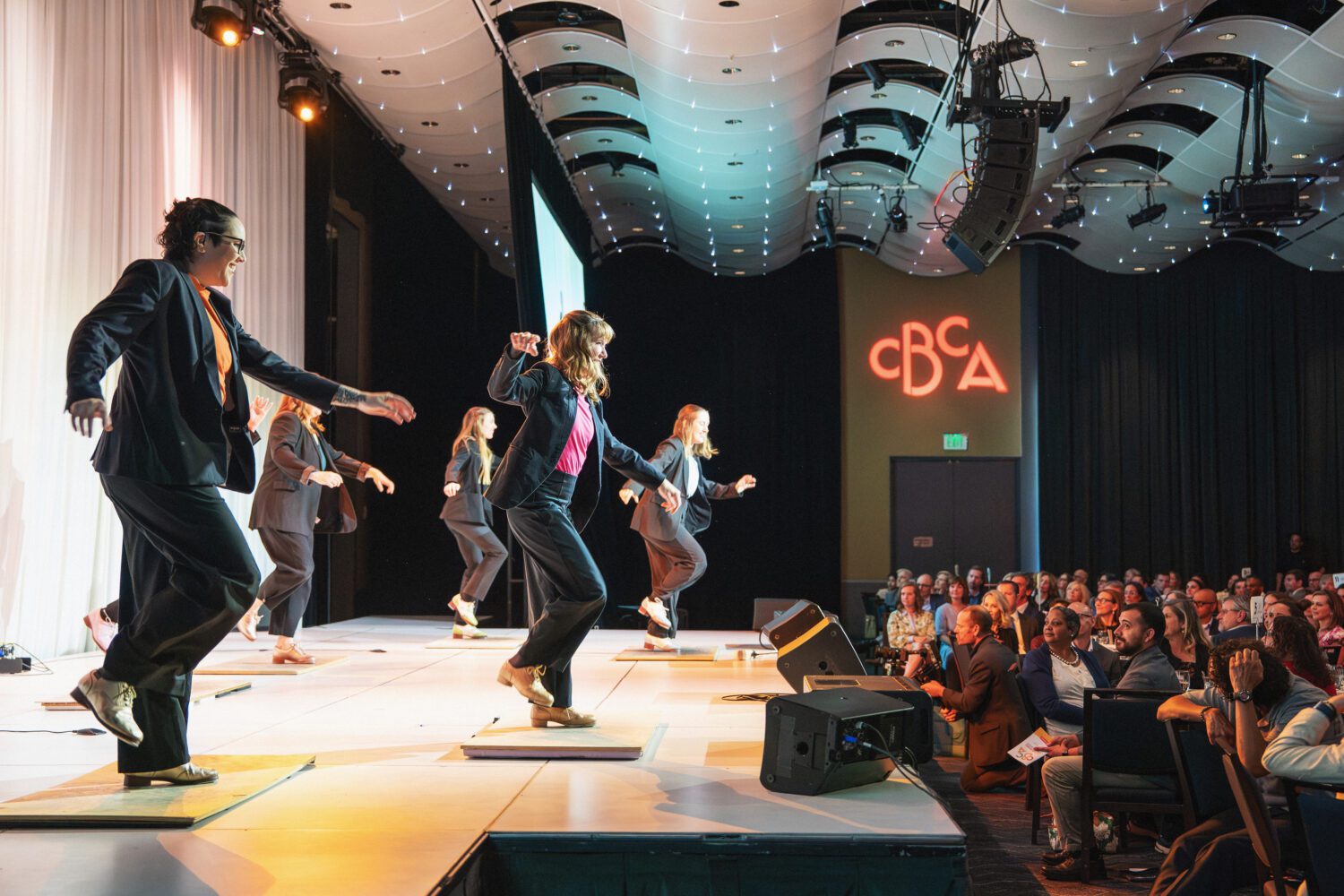 Six people in suits perform a tap dance on stage under bright lights, while an audience watches. A “CBCA” sign glows on the back wall of the darkened theater.