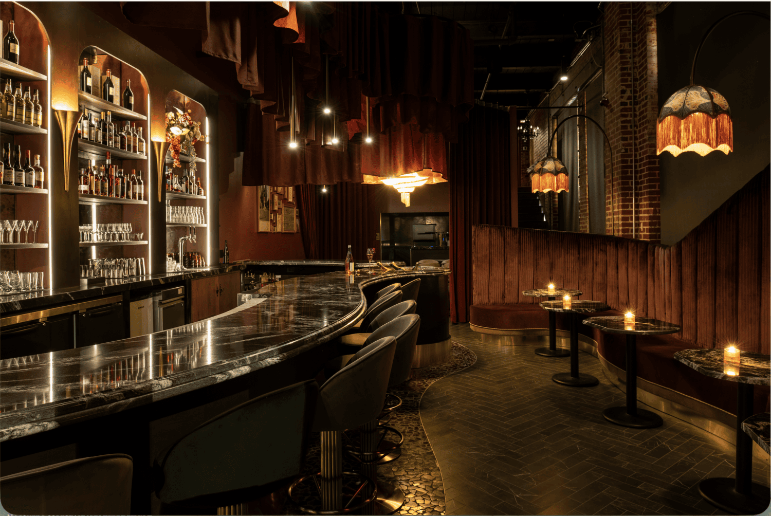 A dimly lit, elegant bar with velvet seating, marble countertops, shelves of bottles, chandeliers, and small candlelit tables, creating a cozy, luxurious atmosphere.